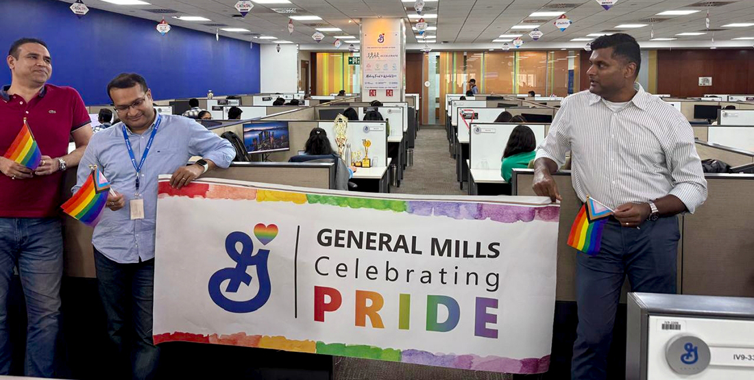 Mini Pride Walk inside the office- A group of people in cubicles holding a banner that reads "General Mills Celebrating Pride" with the General Mills logo displayed.