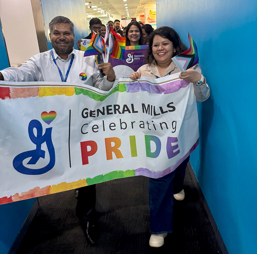Mini Pride Walk inside the office- A group of people holding a rainbow flag that reads "General Mills Celebrating Pride" with the General Mills logo displayed.